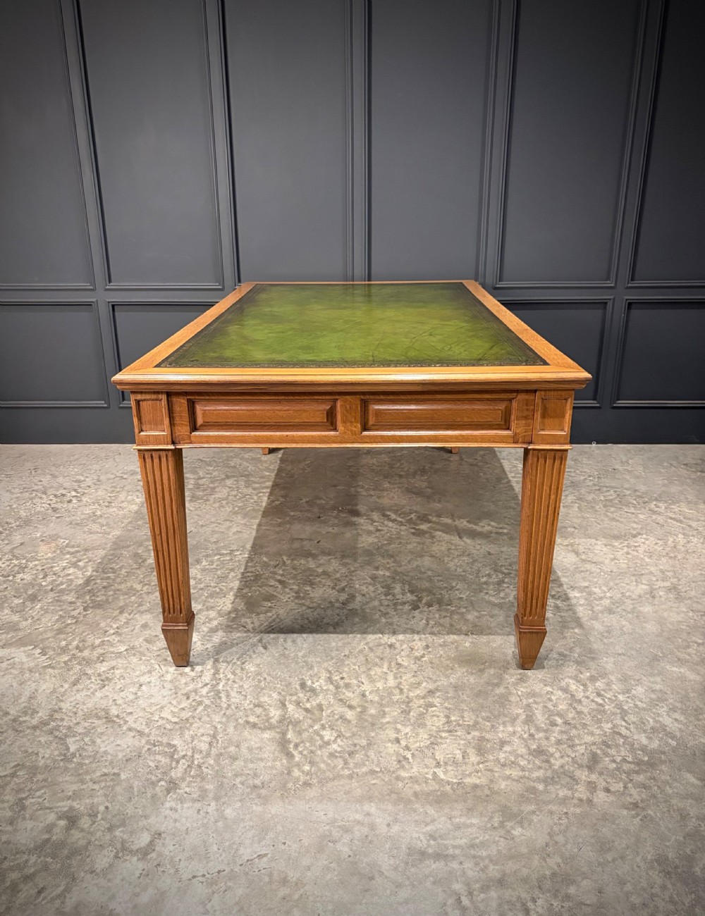 A large oak writing table featuring a green leather top, three frieze drawers with brass drop handles, and four fluted legs. View XIV