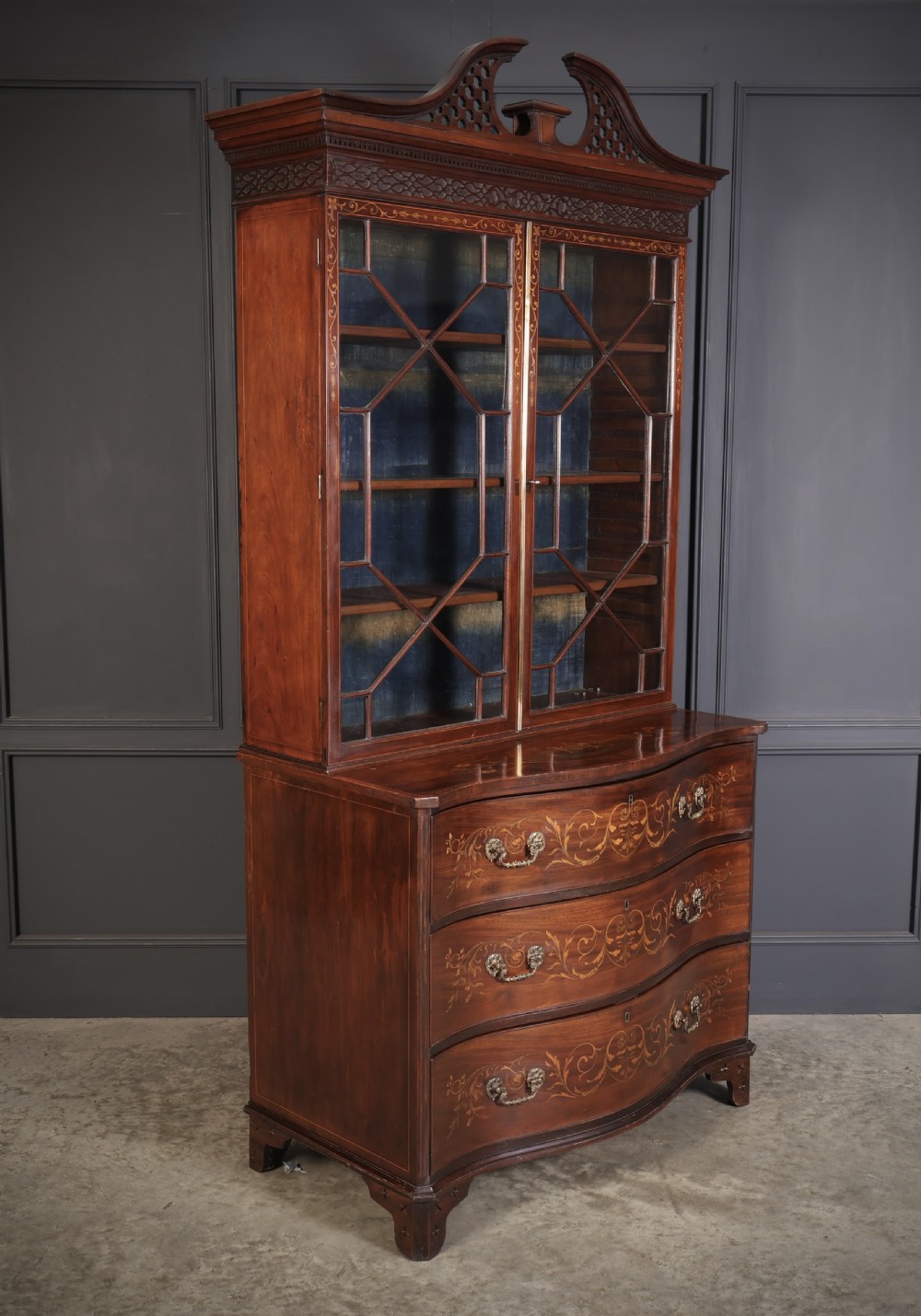 A tall Georgian mahogany bookcase featuring a pierced fretwork pediment, two upper glazed doors with intricate geometric glazing bars, and a lower serpentine chest of three drawers adorned with detailed floral marquetry inlays and brass handles.