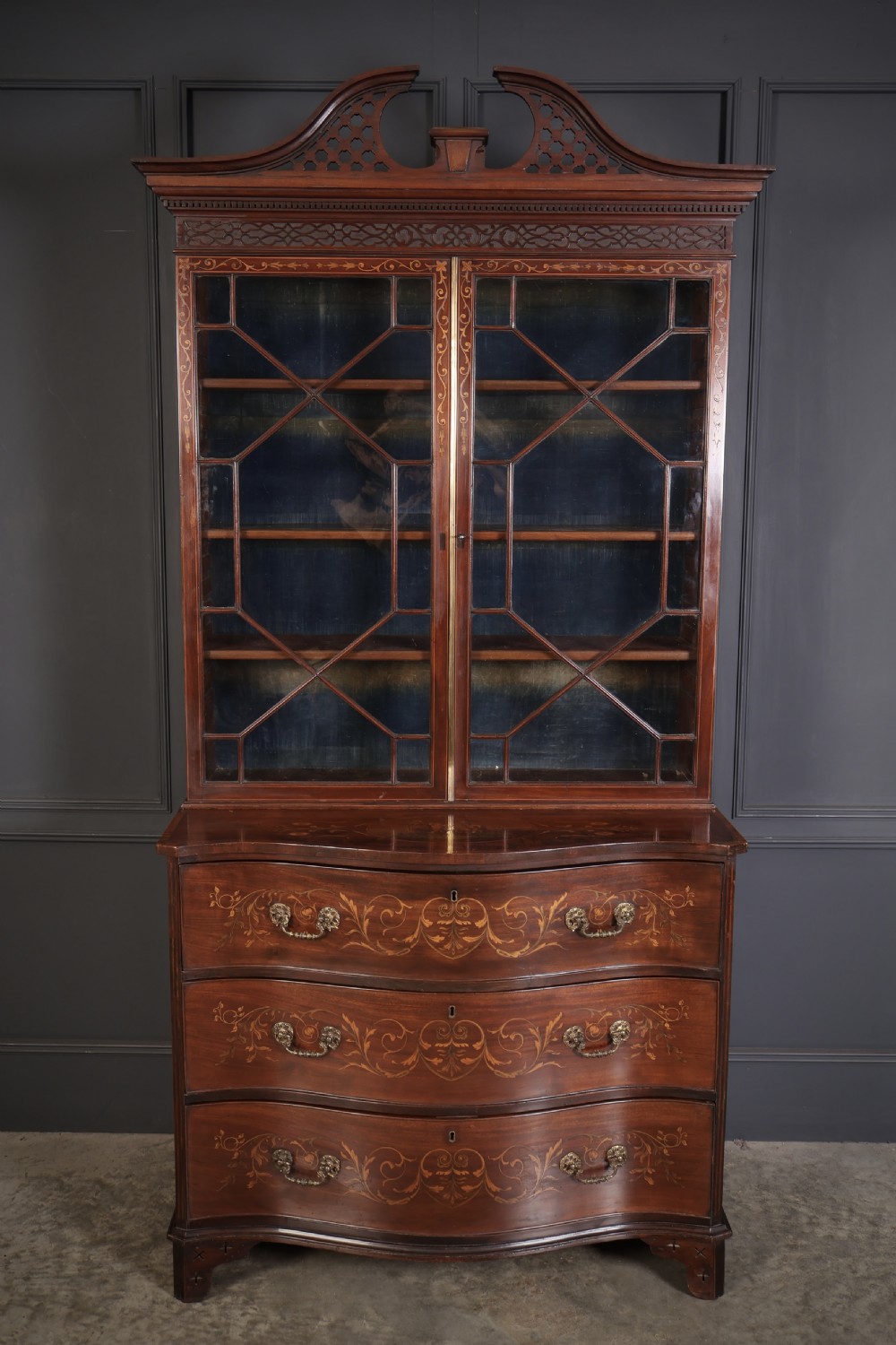 A tall Georgian mahogany bookcase featuring a pierced fretwork pediment, two upper glazed doors with intricate geometric glazing bars, and a lower serpentine chest of three drawers adorned with detailed floral marquetry inlays and brass handles. View VI