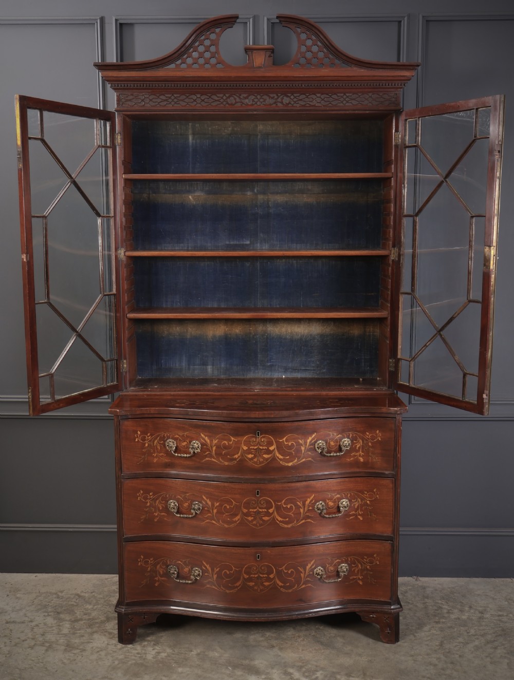 A tall Georgian mahogany bookcase featuring a pierced fretwork pediment, two upper glazed doors with intricate geometric glazing bars, and a lower serpentine chest of three drawers adorned with detailed floral marquetry inlays and brass handles. View VII