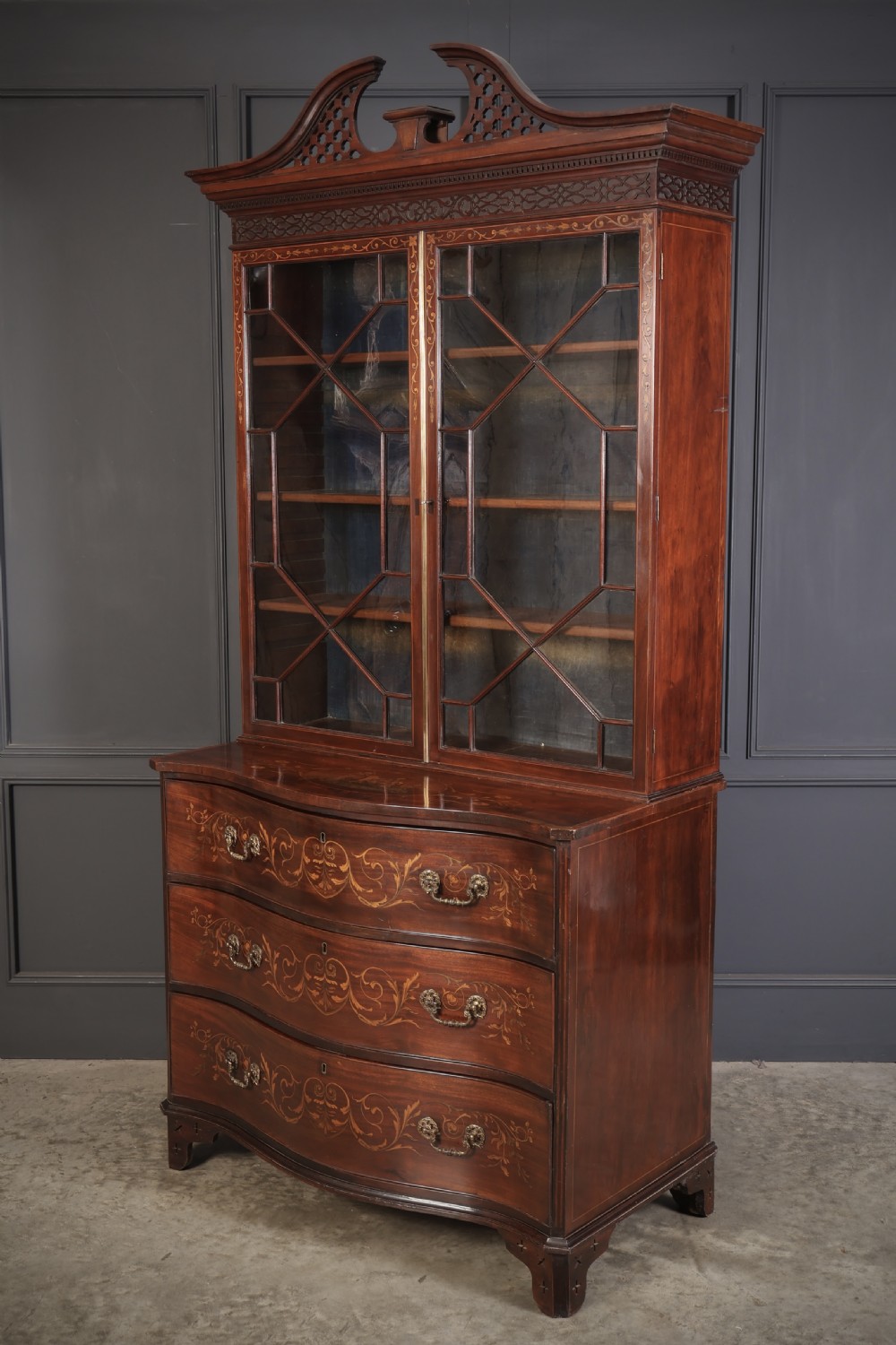 A tall Georgian mahogany bookcase featuring a pierced fretwork pediment, two upper glazed doors with intricate geometric glazing bars, and a lower serpentine chest of three drawers adorned with detailed floral marquetry inlays and brass handles. View IX