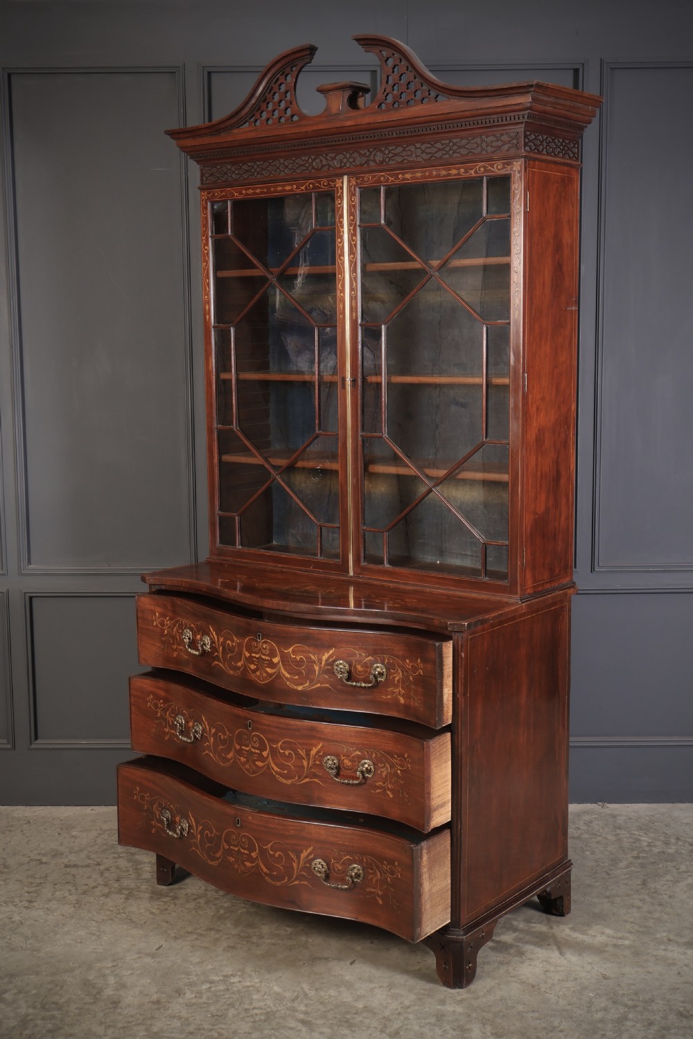 A tall Georgian mahogany bookcase featuring a pierced fretwork pediment, two upper glazed doors with intricate geometric glazing bars, and a lower serpentine chest of three drawers adorned with detailed floral marquetry inlays and brass handles. View XIII