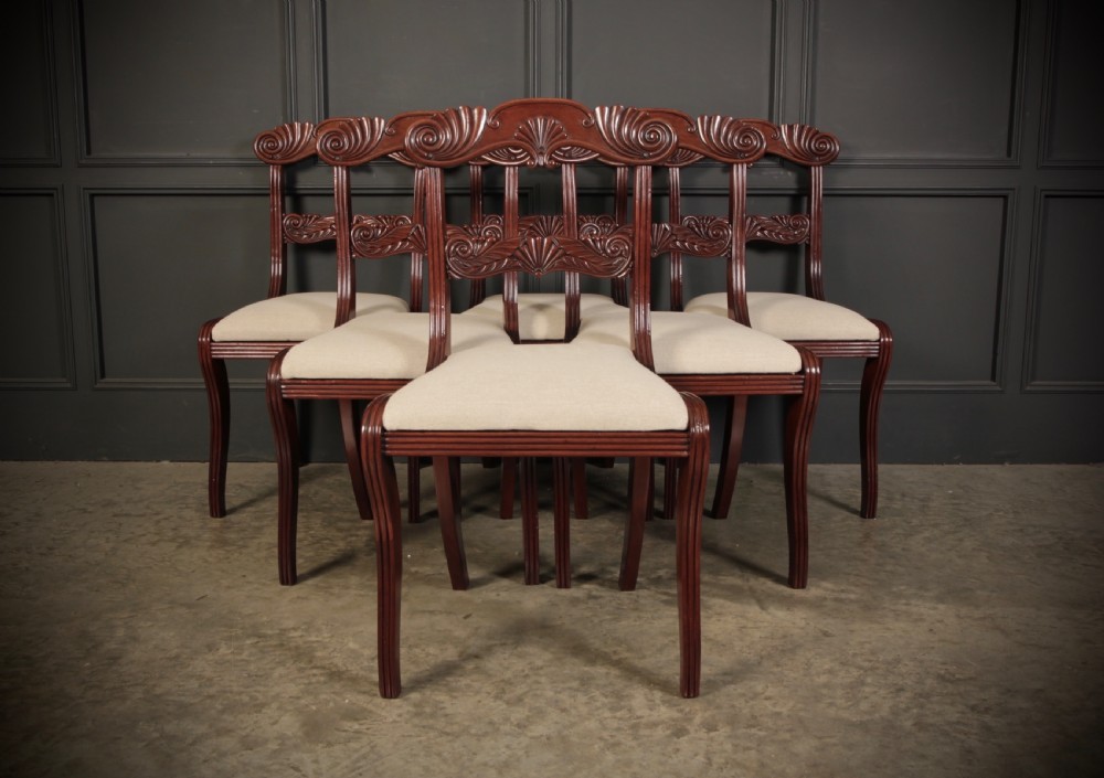 A set of eight carved mahogany Regency dining chairs featuring scroll arms, carved crest rails, and cream upholstered seats. View V