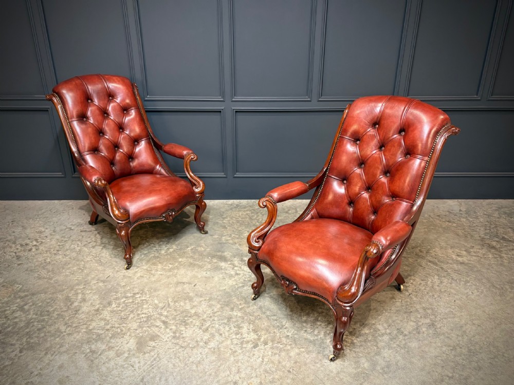 A pair of Victorian library armchairs upholstered in deep-buttoned reddish-brown leather with carved mahogany frames and brass castors.