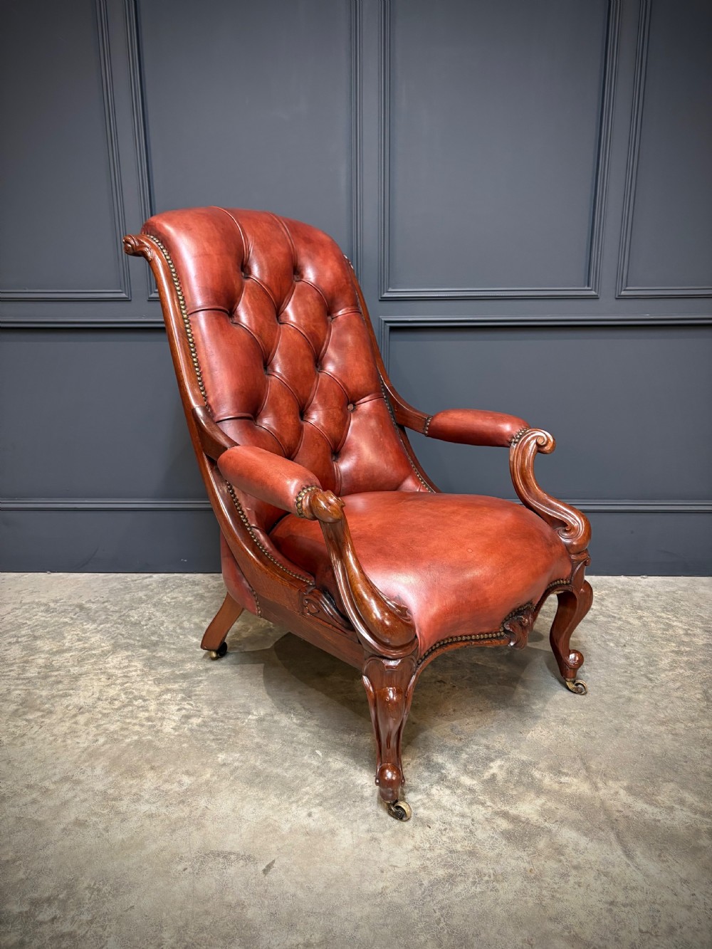 A pair of Victorian library armchairs upholstered in deep-buttoned reddish-brown leather with carved mahogany frames and brass castors. View VI