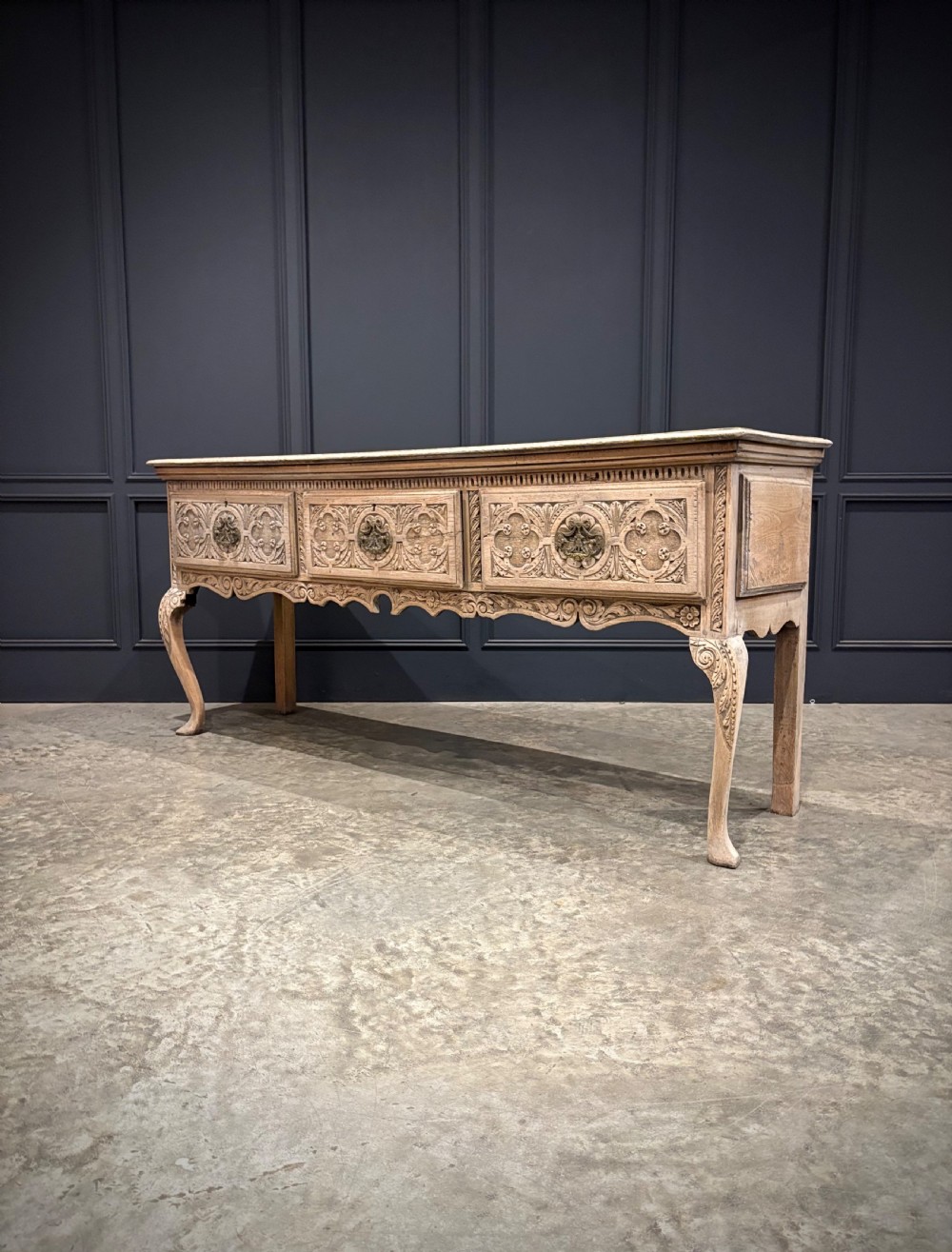 An 18th-century carved oak dresser base featuring three drawers with detailed relief carving, brass handles, shaped apron, cabriole front legs, and a pale bleached finish. View VII