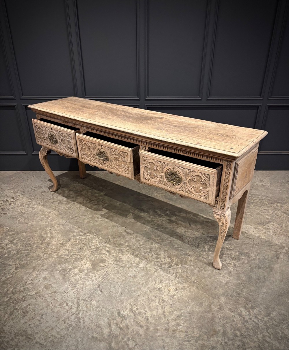 An 18th-century carved oak dresser base featuring three drawers with detailed relief carving, brass handles, shaped apron, cabriole front legs, and a pale bleached finish. View II