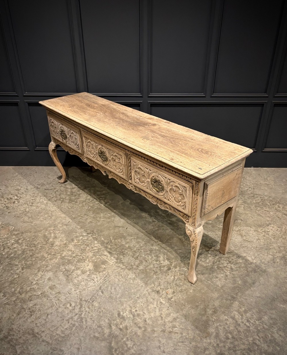 An 18th-century carved oak dresser base featuring three drawers with detailed relief carving, brass handles, shaped apron, cabriole front legs, and a pale bleached finish. View IV