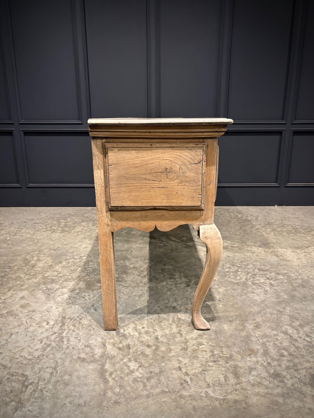 An 18th-century carved oak dresser base featuring three drawers with detailed relief carving, brass handles, shaped apron, cabriole front legs, and a pale bleached finish. View VI