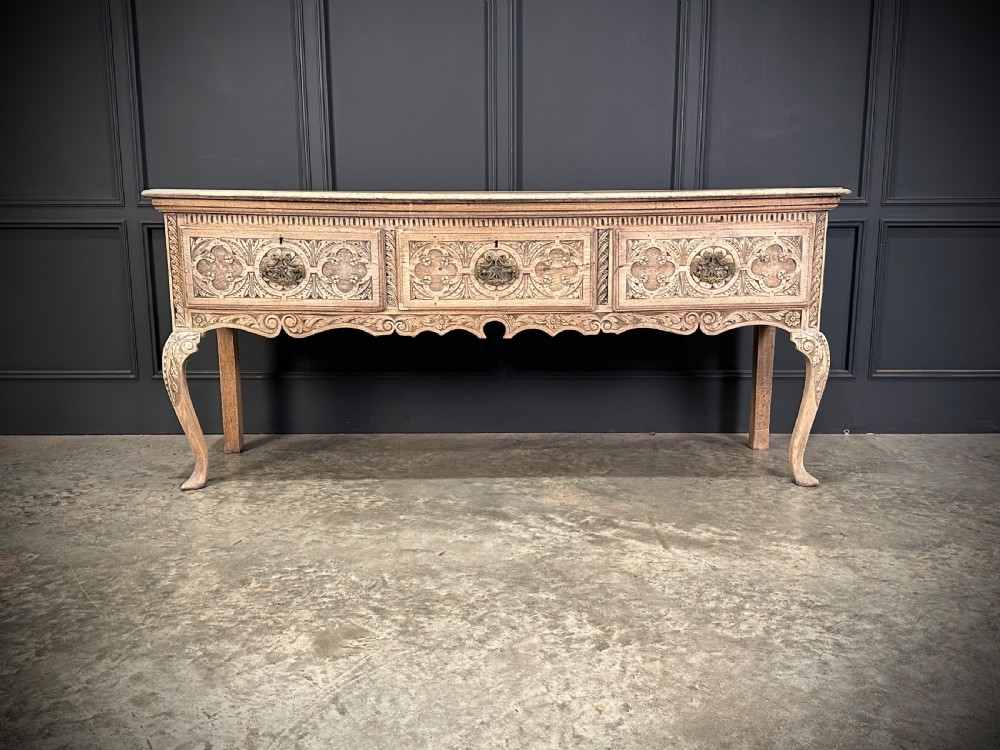An 18th-century carved oak dresser base featuring three drawers with detailed relief carving, brass handles, shaped apron, cabriole front legs, and a pale bleached finish.
