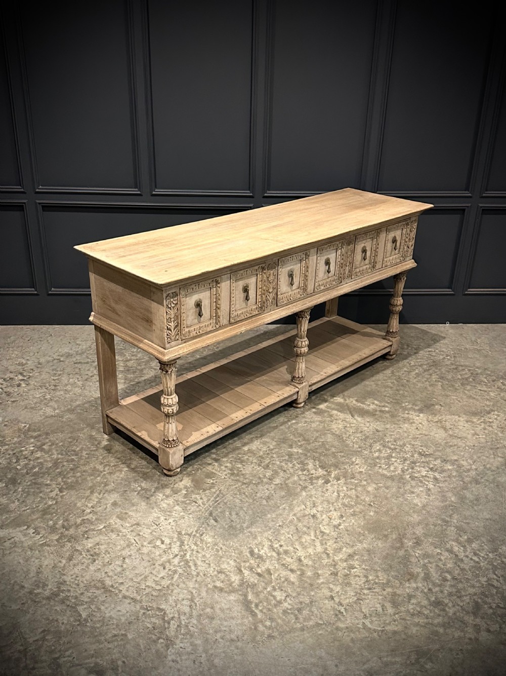 A long carved bleached oak potboard dresser base featuring relief carved drawer fronts, turned support legs, brass drop handles, and an open lower display shelf. View VI