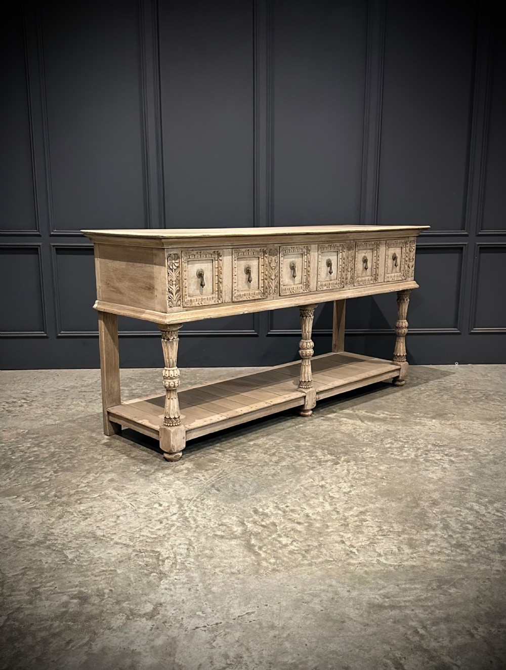 A long carved bleached oak potboard dresser base featuring relief carved drawer fronts, turned support legs, brass drop handles, and an open lower display shelf. View II