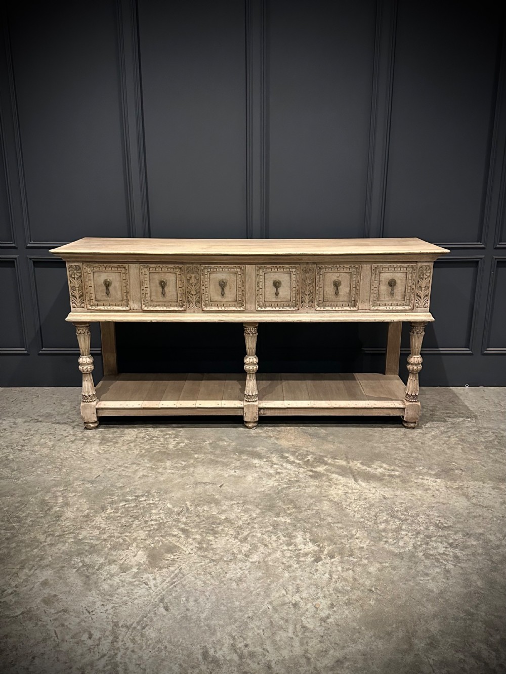 A long carved bleached oak potboard dresser base featuring relief carved drawer fronts, turned support legs, brass drop handles, and an open lower display shelf. View V