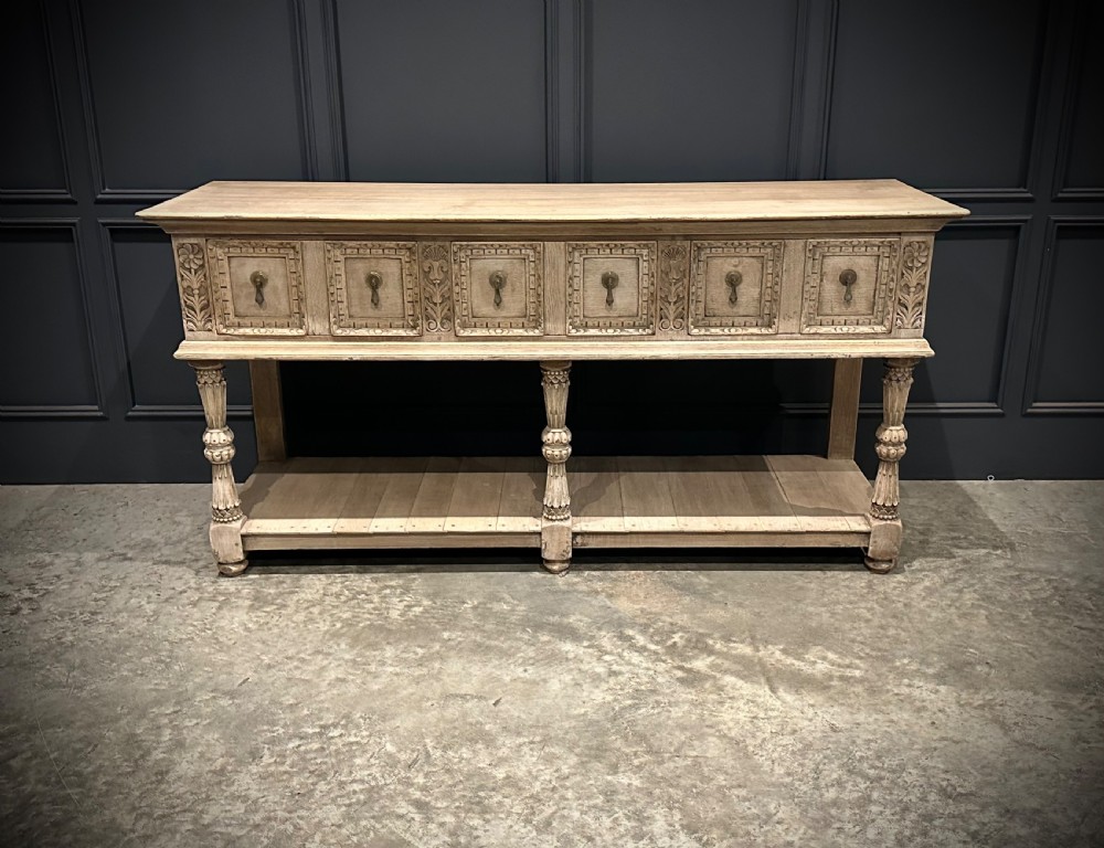 A long carved bleached oak potboard dresser base featuring relief carved drawer fronts, turned support legs, brass drop handles, and an open lower display shelf.