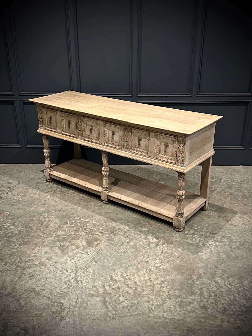 A long carved bleached oak potboard dresser base featuring relief carved drawer fronts, turned support legs, brass drop handles, and an open lower display shelf. View VIII
