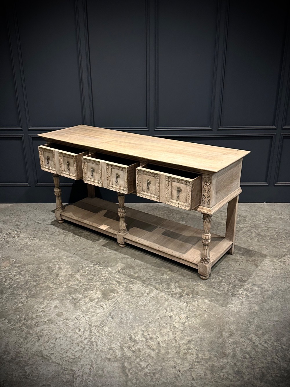 A long carved bleached oak potboard dresser base featuring relief carved drawer fronts, turned support legs, brass drop handles, and an open lower display shelf. View XI