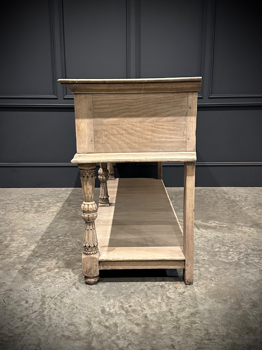 A long carved bleached oak potboard dresser base featuring relief carved drawer fronts, turned support legs, brass drop handles, and an open lower display shelf. View XII