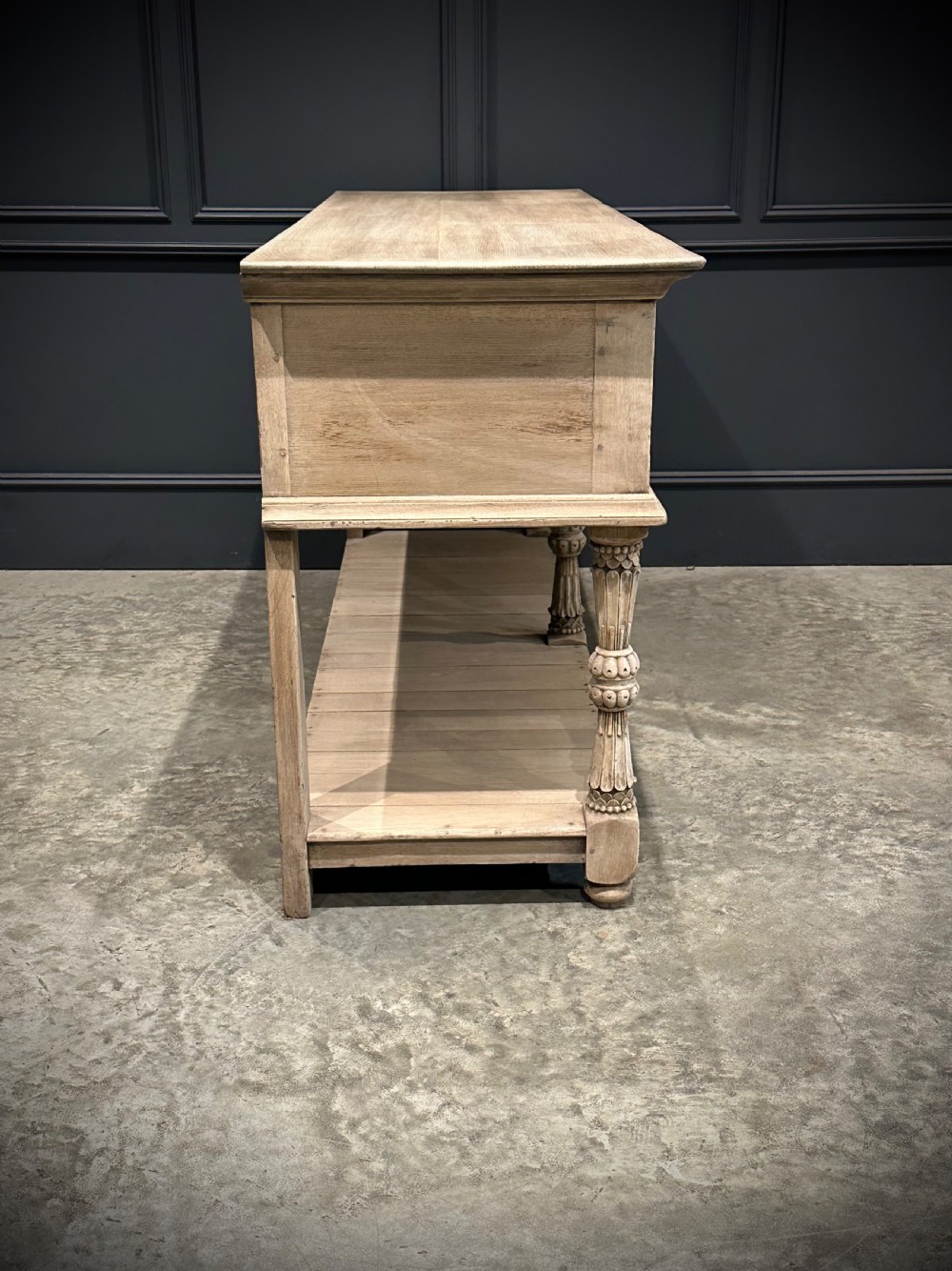 A long carved bleached oak potboard dresser base featuring relief carved drawer fronts, turned support legs, brass drop handles, and an open lower display shelf. View XIV