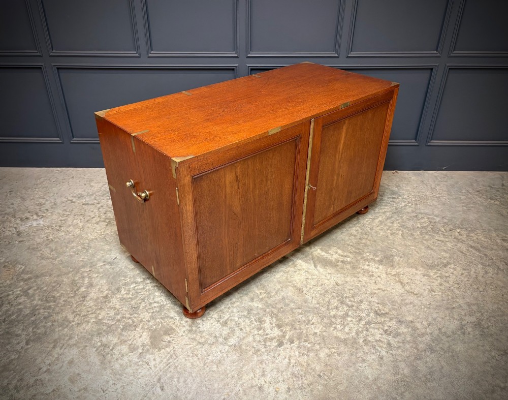 A low Victorian teak military campaign side cabinet featuring double panelled doors, brass corner mounts, brass side handles, and short turned feet. View II