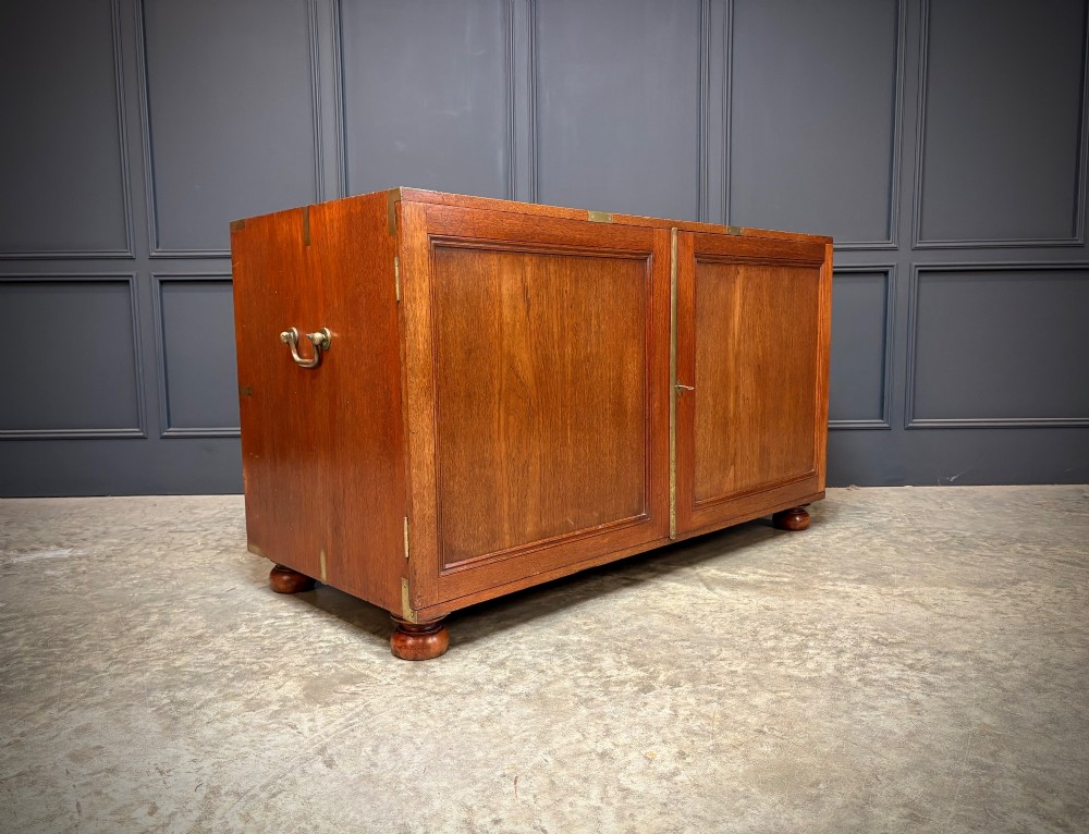 A low Victorian teak military campaign side cabinet featuring double panelled doors, brass corner mounts, brass side handles, and short turned feet.