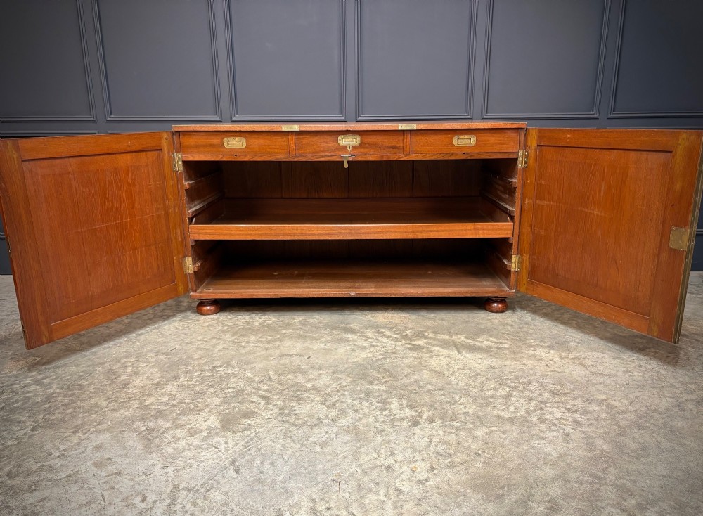 A low Victorian teak military campaign side cabinet featuring double panelled doors, brass corner mounts, brass side handles, and short turned feet. View V