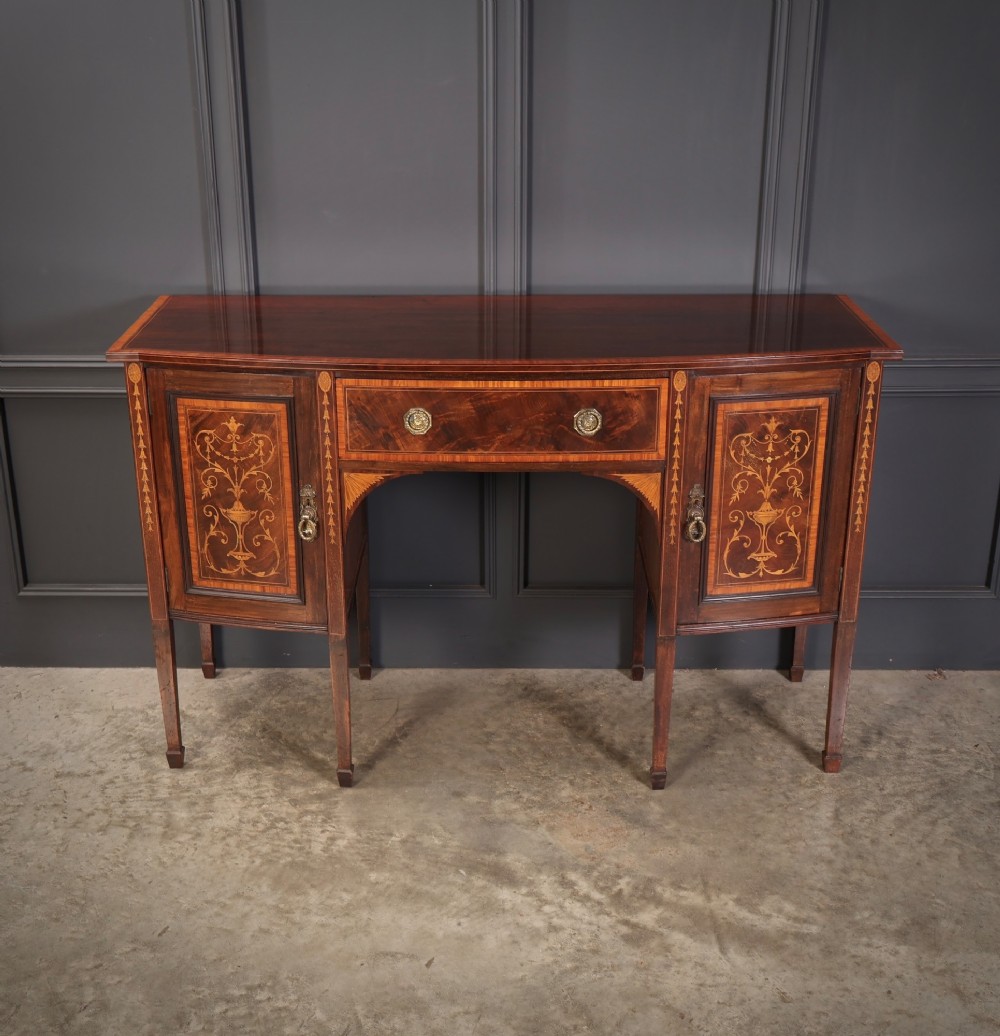An antique bowfront mahogany sideboard featuring intricate urn marquetry inlays on cupboard doors, a central drawer, and slender square tapered legs.