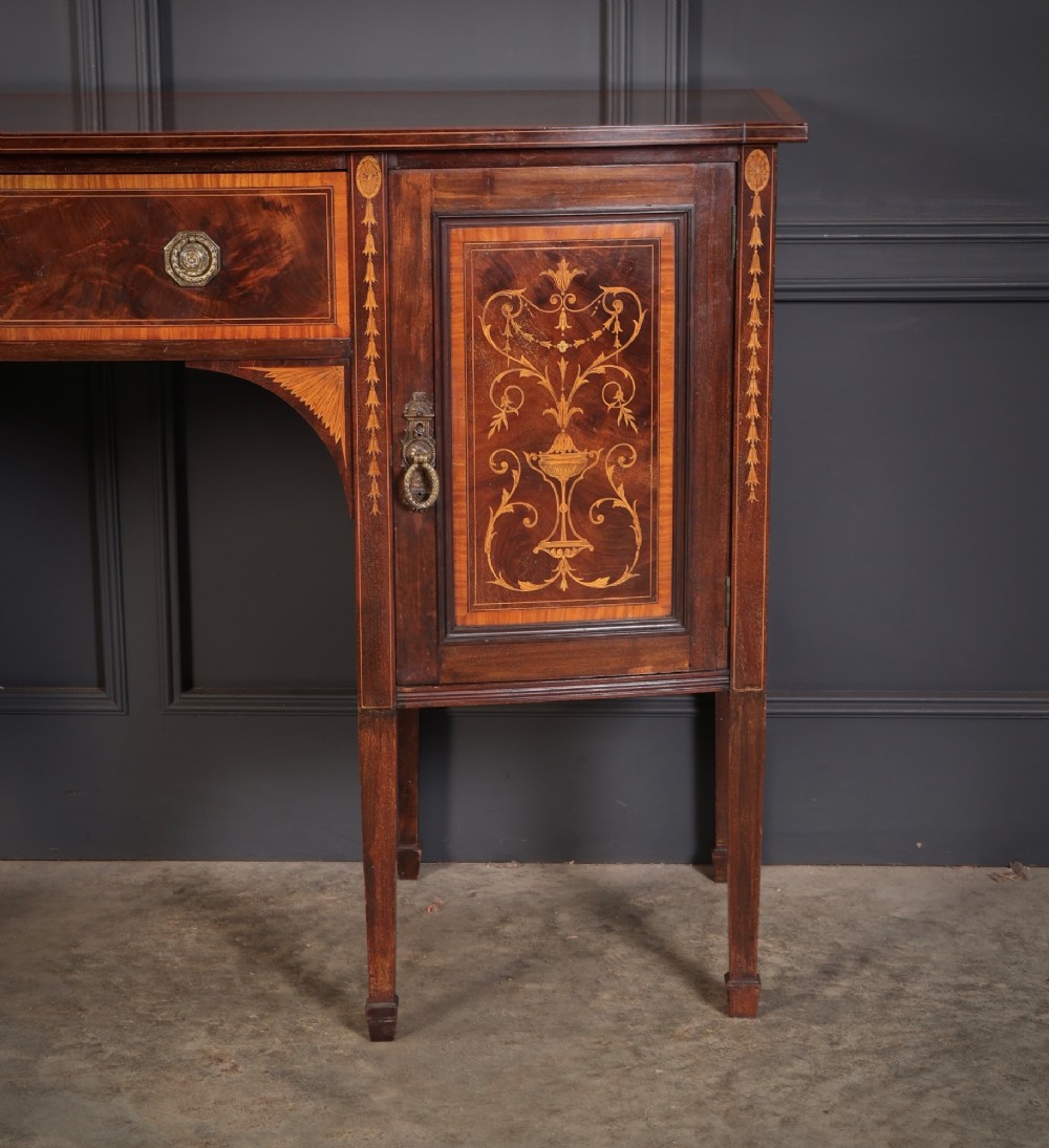 An antique bowfront mahogany sideboard featuring intricate urn marquetry inlays on cupboard doors, a central drawer, and slender square tapered legs. View III