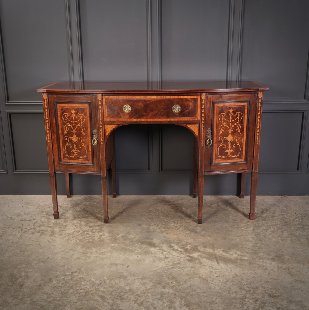 An antique bowfront mahogany sideboard featuring intricate urn marquetry inlays on cupboard doors, a central drawer, and slender square tapered legs. View IV