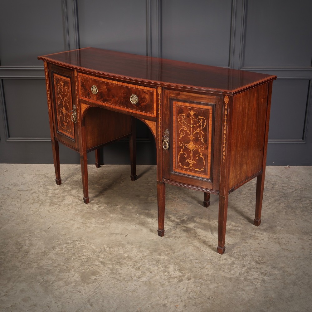 An antique bowfront mahogany sideboard featuring intricate urn marquetry inlays on cupboard doors, a central drawer, and slender square tapered legs. View V