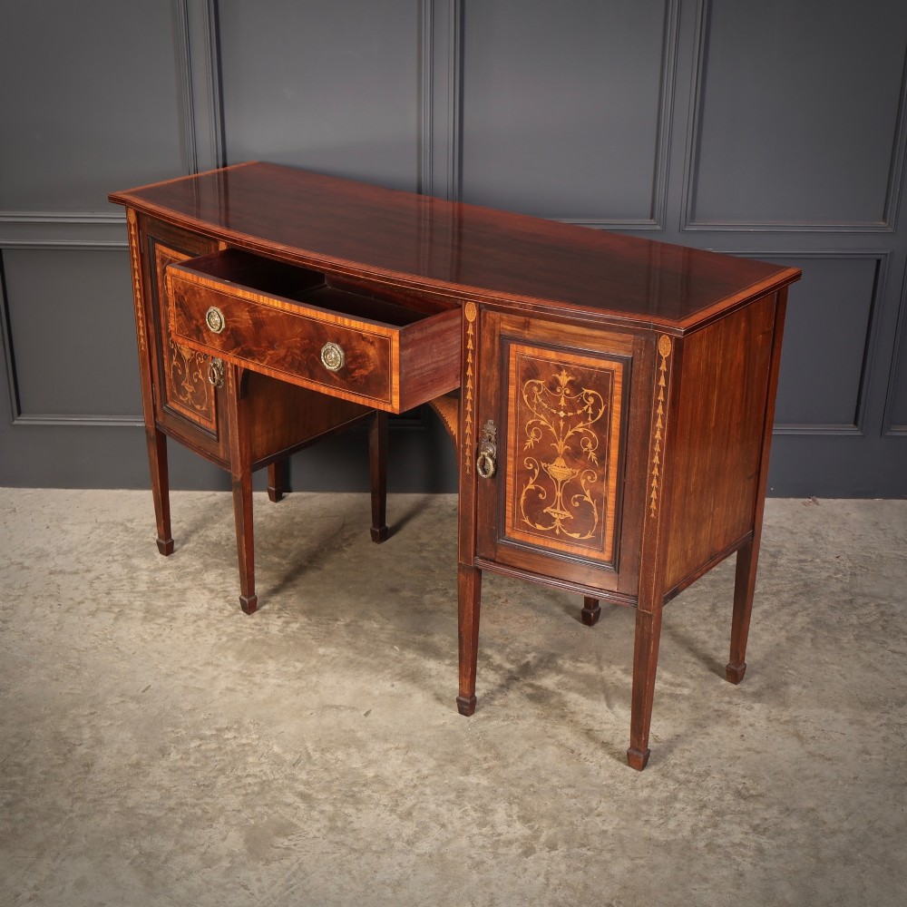 An antique bowfront mahogany sideboard featuring intricate urn marquetry inlays on cupboard doors, a central drawer, and slender square tapered legs. View VI