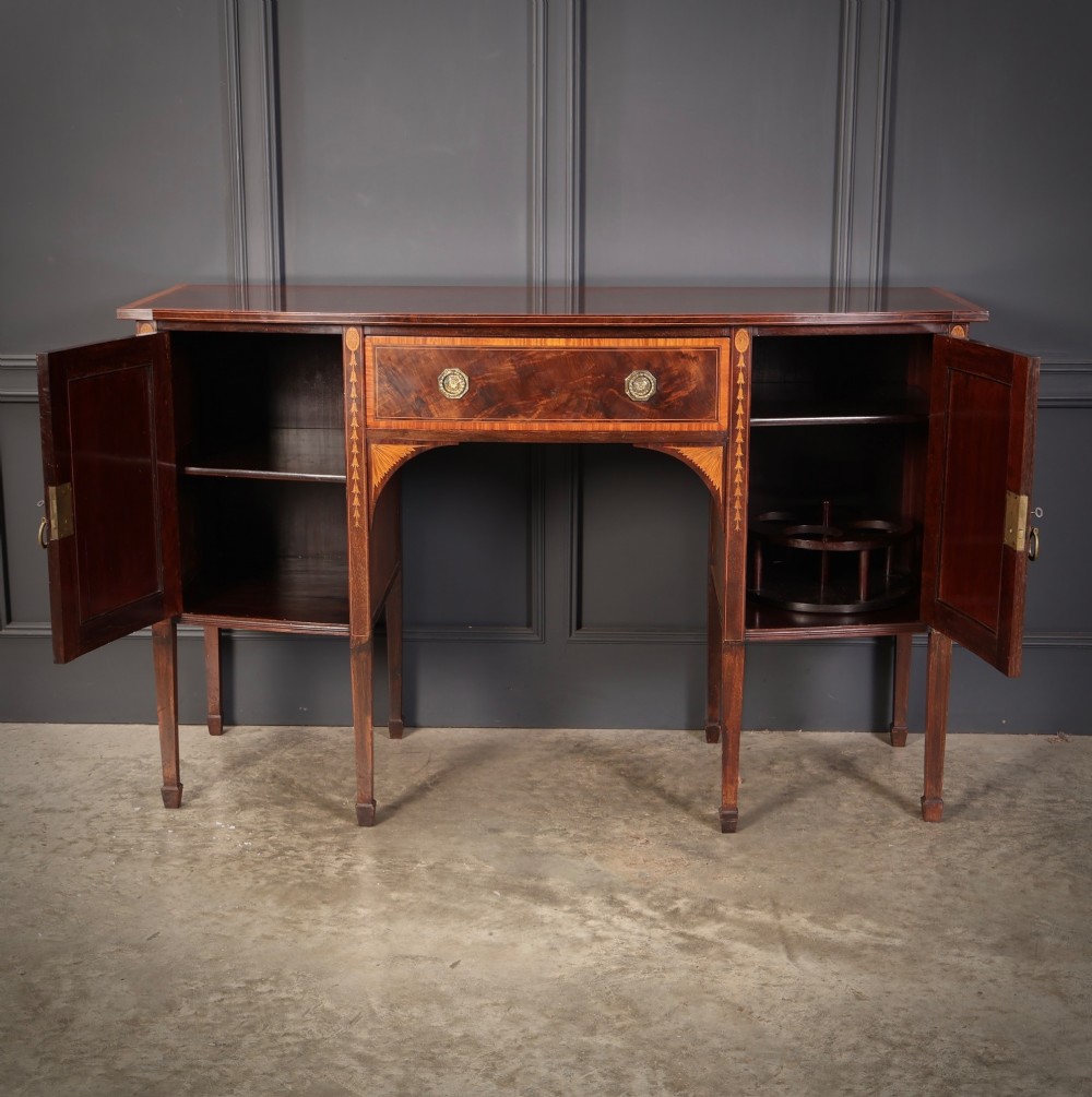 An antique bowfront mahogany sideboard featuring intricate urn marquetry inlays on cupboard doors, a central drawer, and slender square tapered legs. View VII