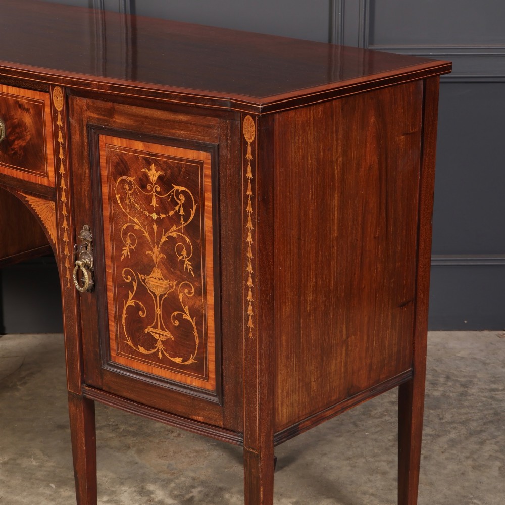 An antique bowfront mahogany sideboard featuring intricate urn marquetry inlays on cupboard doors, a central drawer, and slender square tapered legs. View X
