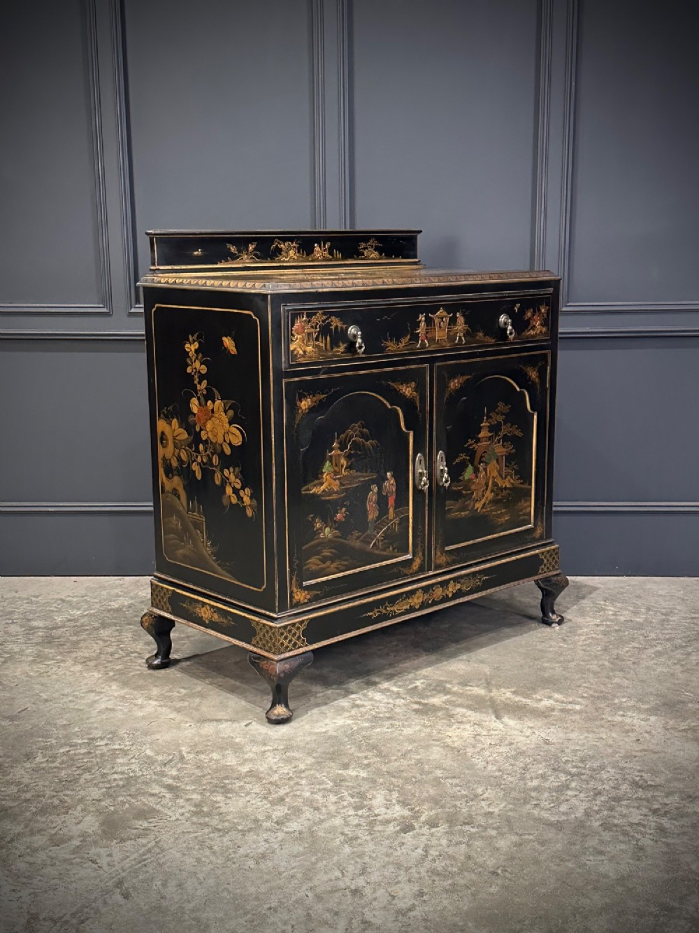 A black lacquered side cabinet decorated with gold chinoiserie landscape scenes, featuring one top drawer, double doors, and cabriole legs.
