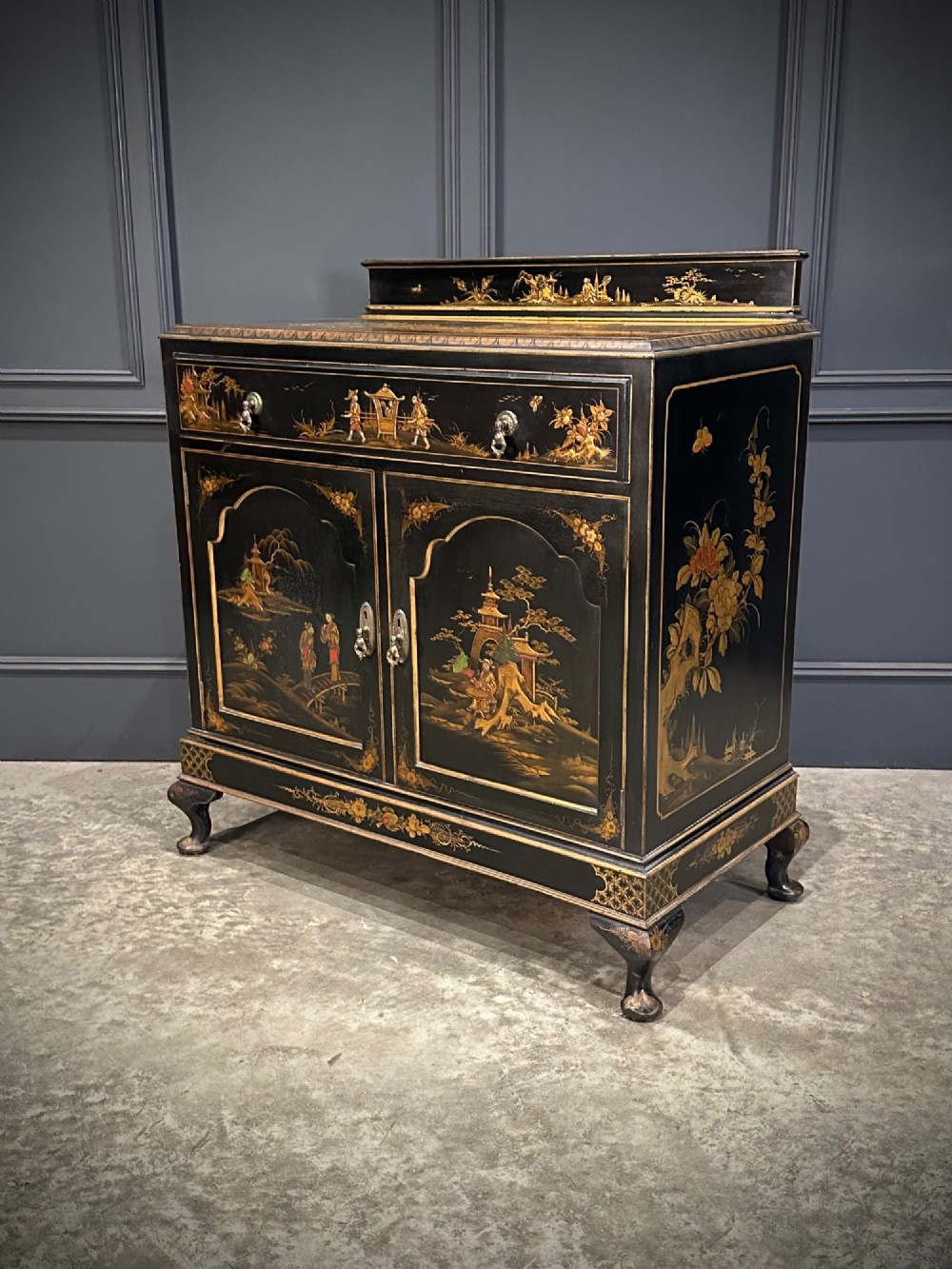 A black lacquered side cabinet decorated with gold chinoiserie landscape scenes, featuring one top drawer, double doors, and cabriole legs. View IX