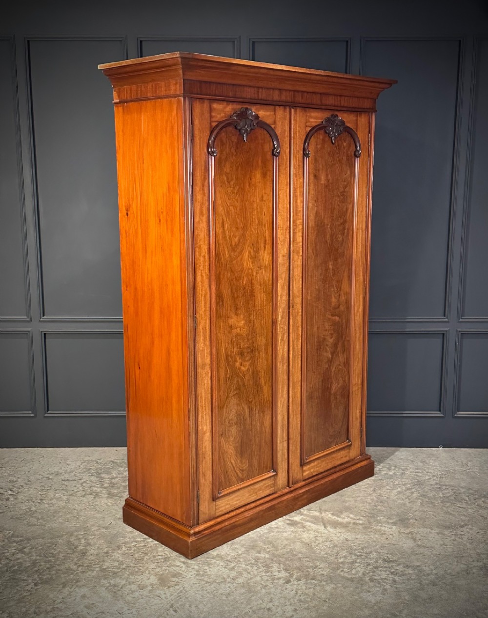 A Victorian mahogany double wardrobe featuring two arched panel doors with carved crest details, a flared top cornice, and a solid plinth base.