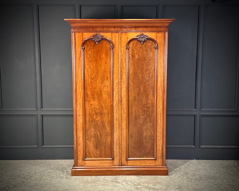 A Victorian mahogany double wardrobe featuring two arched panel doors with carved crest details, a flared top cornice, and a solid plinth base. View II