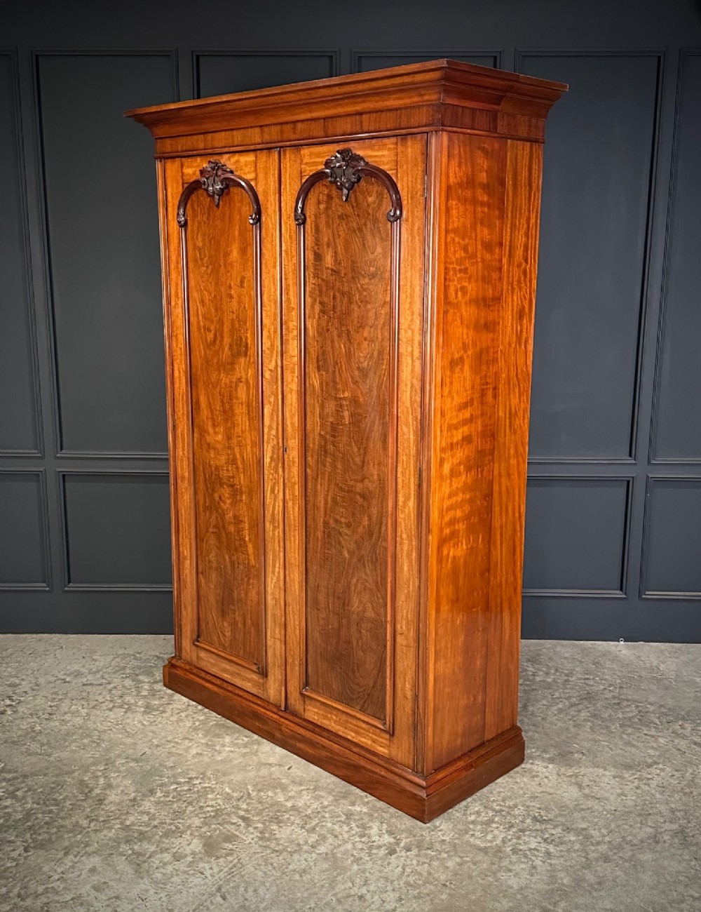 A Victorian mahogany double wardrobe featuring two arched panel doors with carved crest details, a flared top cornice, and a solid plinth base. View VII