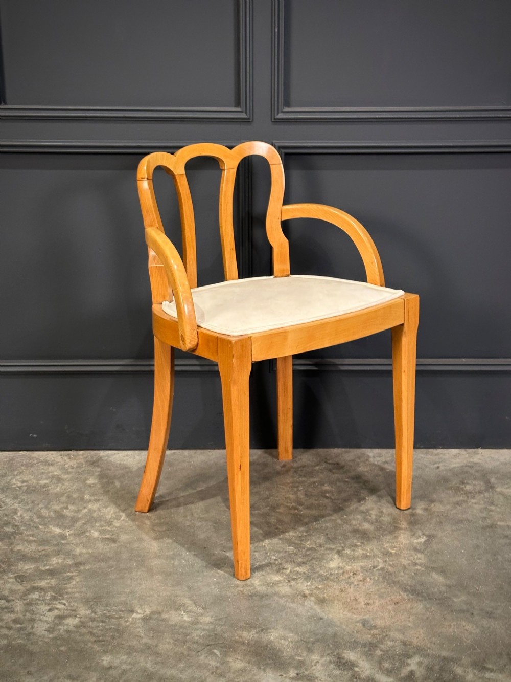 An Art Deco satinwood armchair featuring a triple-arched back rest, curved sweeping arms, a light cream upholstered seat pad, and tapered legs.