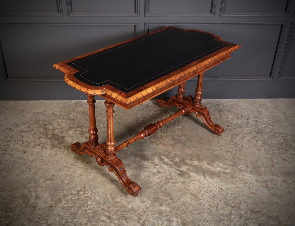 A carved walnut library table featuring a dark leather inset writing surface, detailed apron moulding, turned twin supports, and a carved trestle base with scroll feet. View II