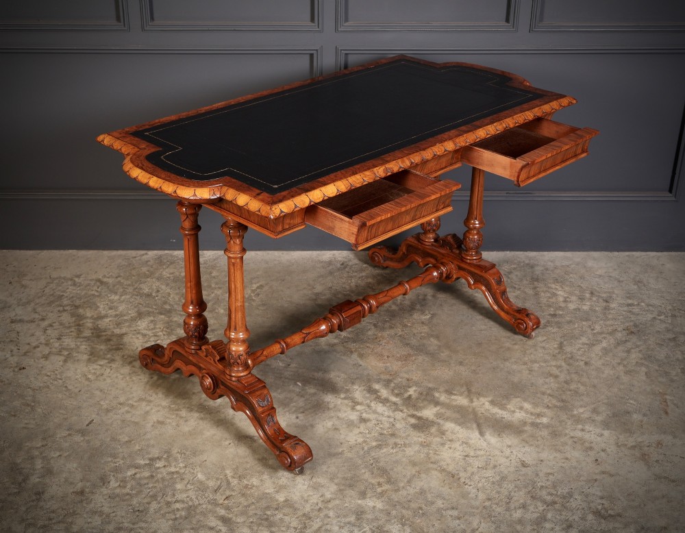A carved walnut library table featuring a dark leather inset writing surface, detailed apron moulding, turned twin supports, and a carved trestle base with scroll feet. View III