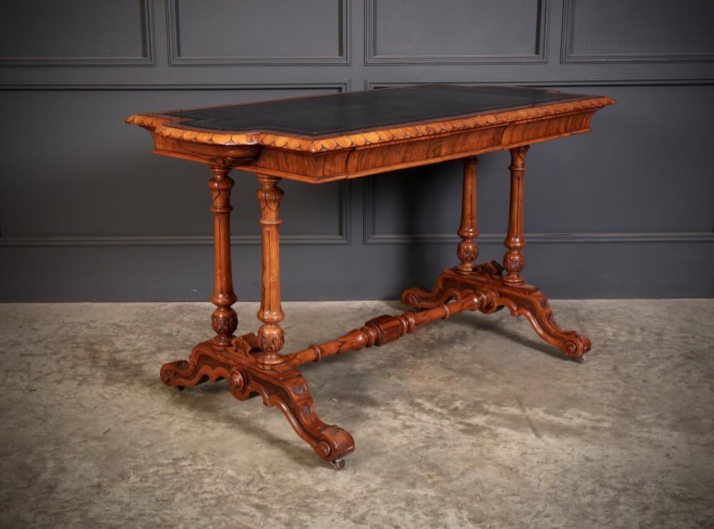 A carved walnut library table featuring a dark leather inset writing surface, detailed apron moulding, turned twin supports, and a carved trestle base with scroll feet.