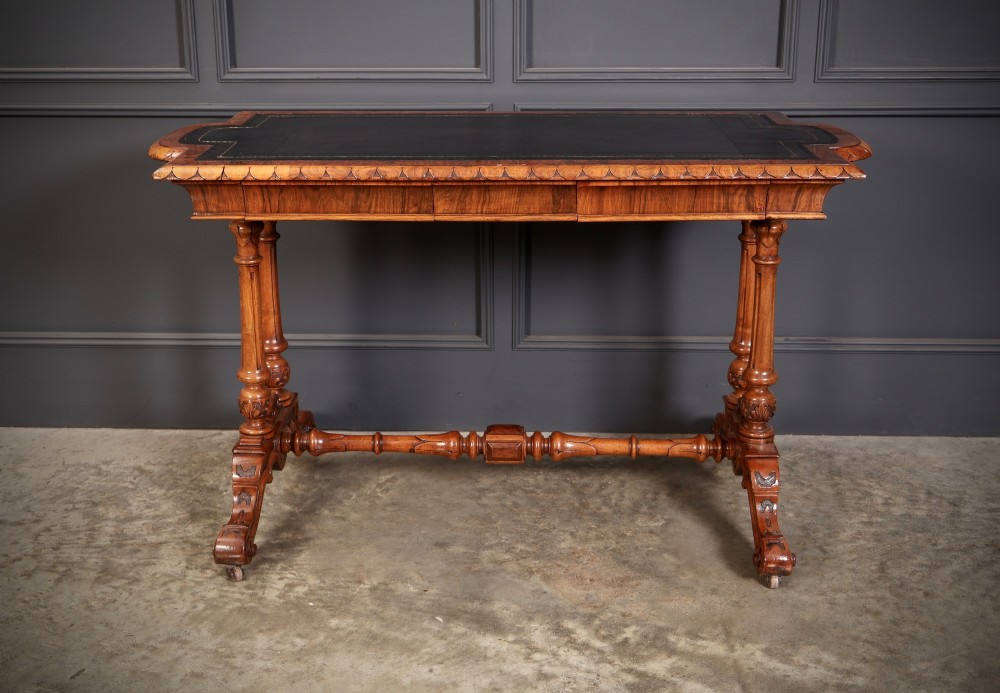 A carved walnut library table featuring a dark leather inset writing surface, detailed apron moulding, turned twin supports, and a carved trestle base with scroll feet. View V