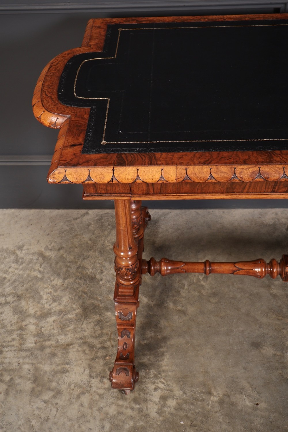 A carved walnut library table featuring a dark leather inset writing surface, detailed apron moulding, turned twin supports, and a carved trestle base with scroll feet. View VI