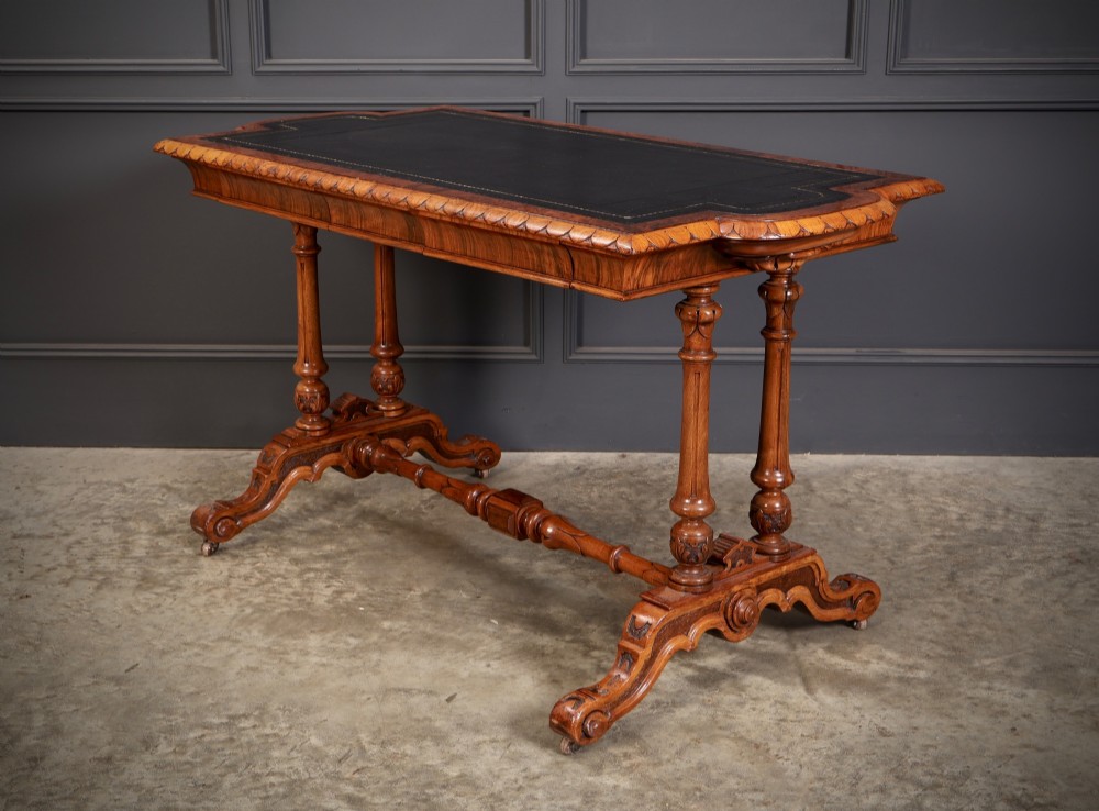 A carved walnut library table featuring a dark leather inset writing surface, detailed apron moulding, turned twin supports, and a carved trestle base with scroll feet. View VII