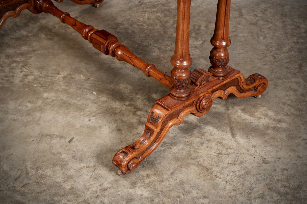 A carved walnut library table featuring a dark leather inset writing surface, detailed apron moulding, turned twin supports, and a carved trestle base with scroll feet. View VIII