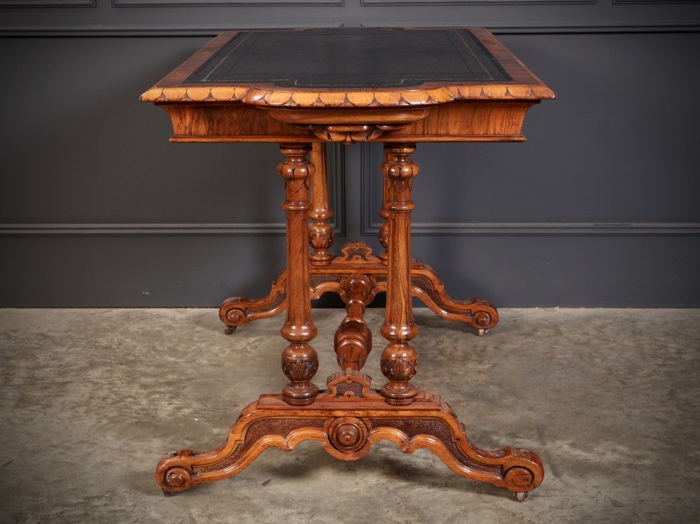 A carved walnut library table featuring a dark leather inset writing surface, detailed apron moulding, turned twin supports, and a carved trestle base with scroll feet. View IX