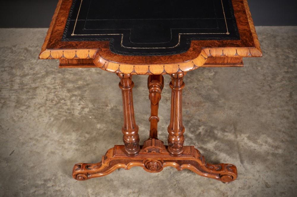 A carved walnut library table featuring a dark leather inset writing surface, detailed apron moulding, turned twin supports, and a carved trestle base with scroll feet. View X