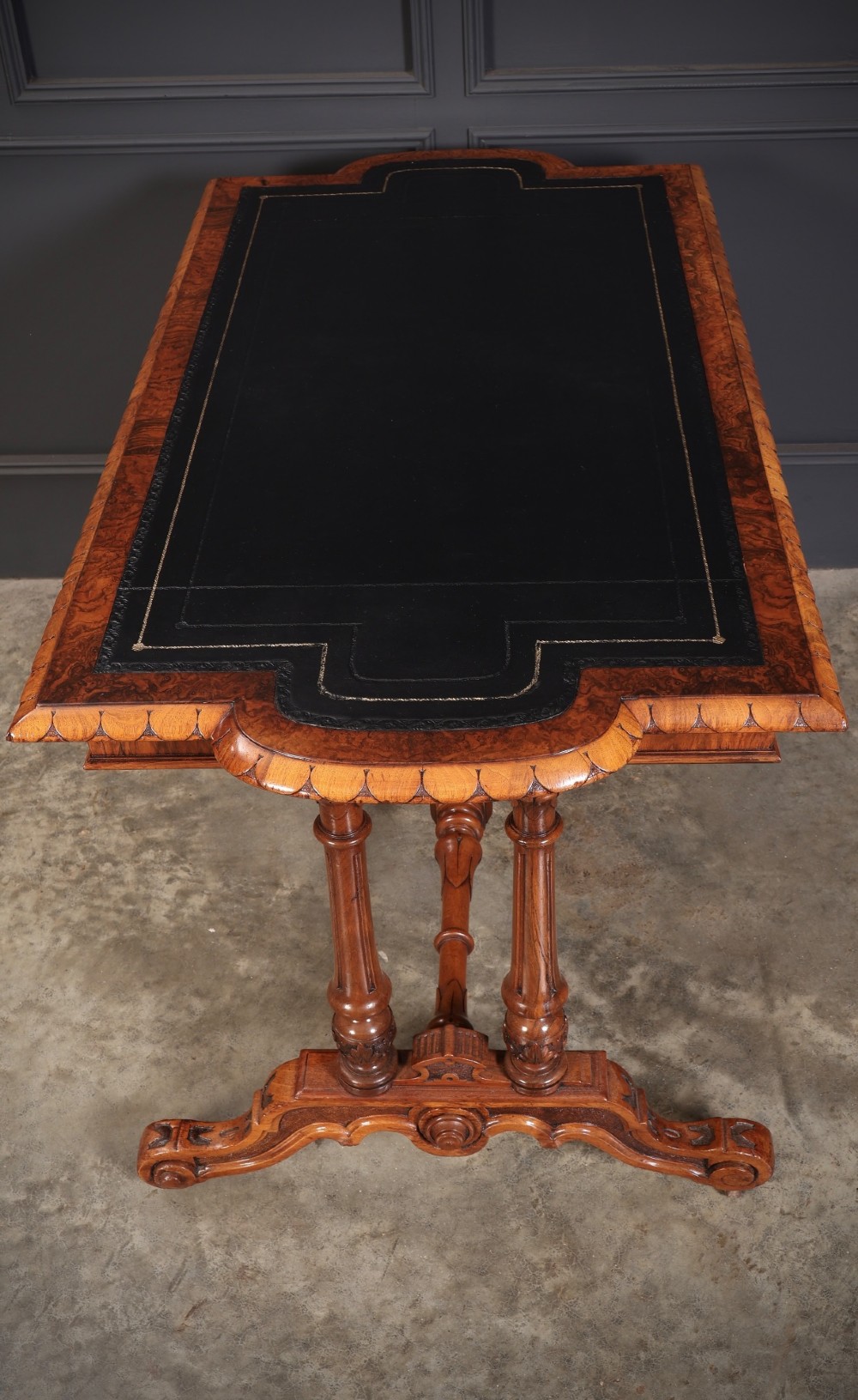A carved walnut library table featuring a dark leather inset writing surface, detailed apron moulding, turned twin supports, and a carved trestle base with scroll feet. View XI