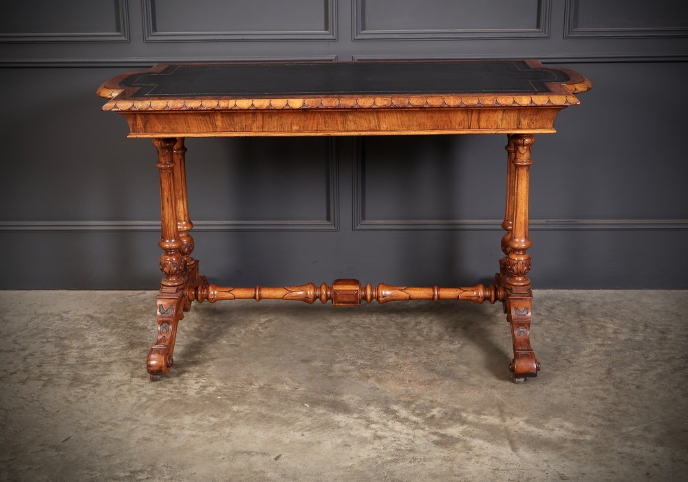 A carved walnut library table featuring a dark leather inset writing surface, detailed apron moulding, turned twin supports, and a carved trestle base with scroll feet. View XII