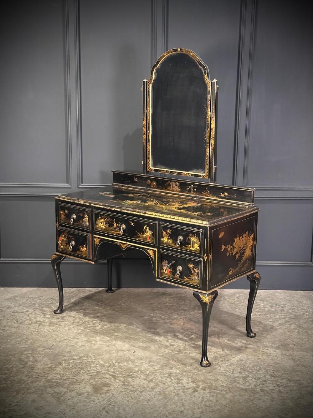 A black lacquered Chinoiserie dressing table featuring gilt hand-painted figures and landscapes, five drawers, cabriole legs, and an arched mirror attached to the top.